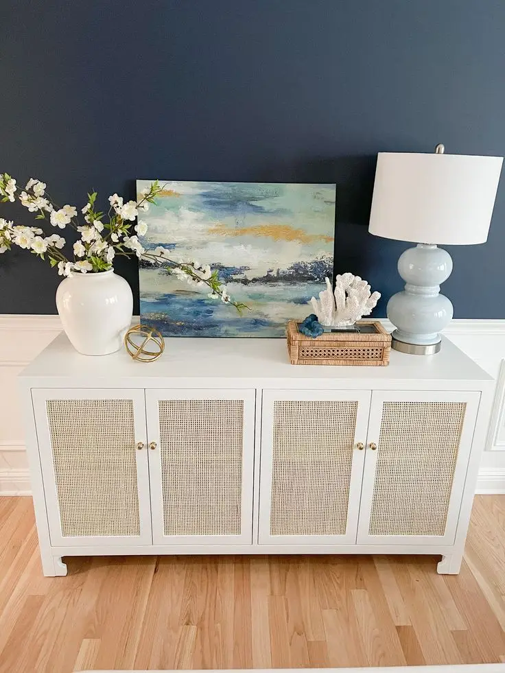 A cane door cabinet with a beach painting, a plant and a lamp.