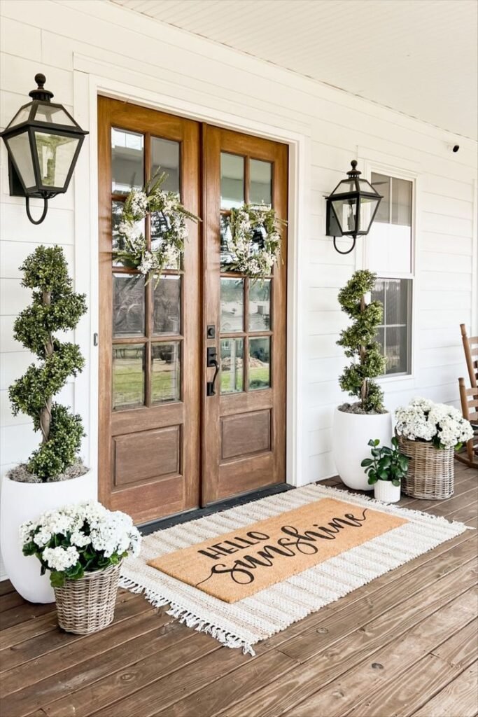 A big wooden door with two summer wreaths, flowers and a welcome mat.
