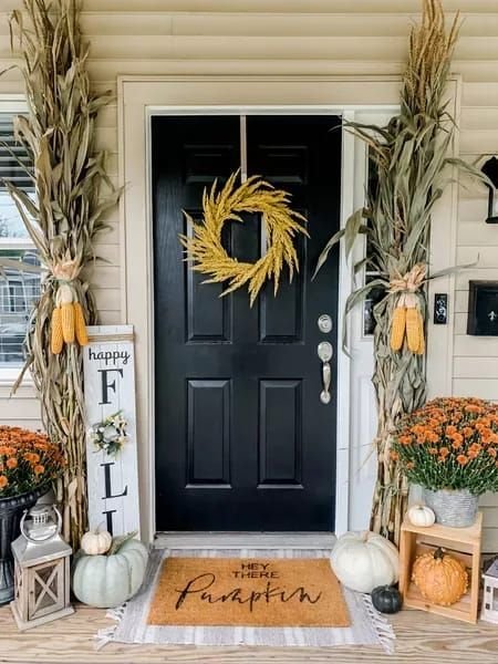 A black door with a dried plants wreath. This front porch has a fall wooden sign, chrysanthemums and cornstalks.