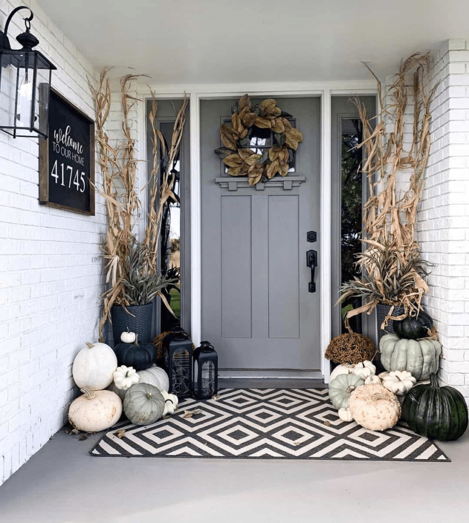 A gray door with a fall wreath, geometrical rug, cornstalks and many pumpkins of different shapes, sizes and colors.