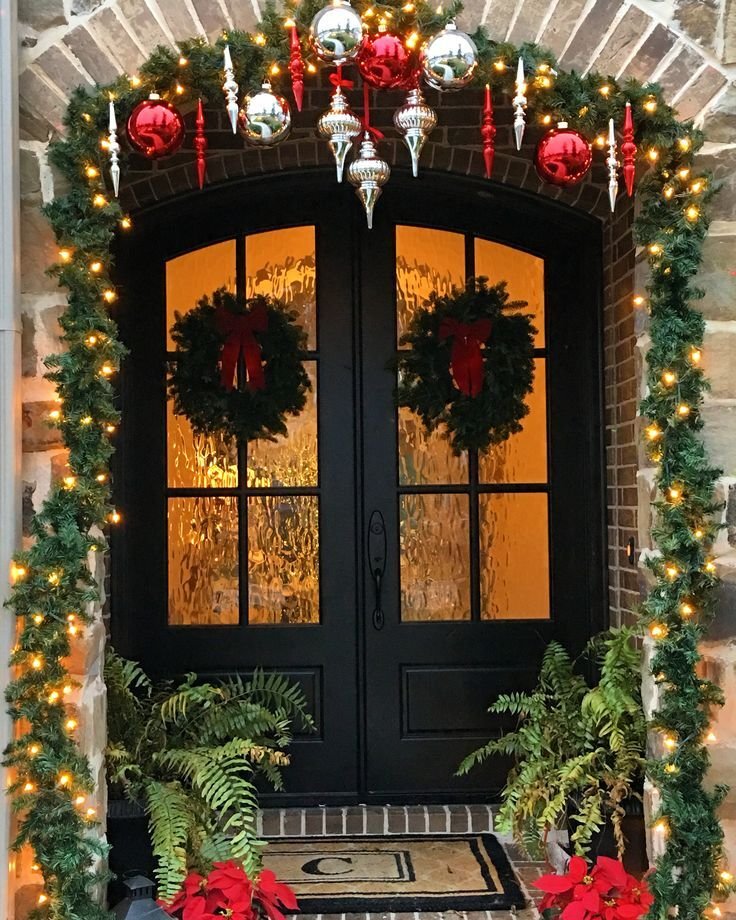 A big black door with two Christmas wreaths, a garland with string lights and ornaments hanging from the wall.