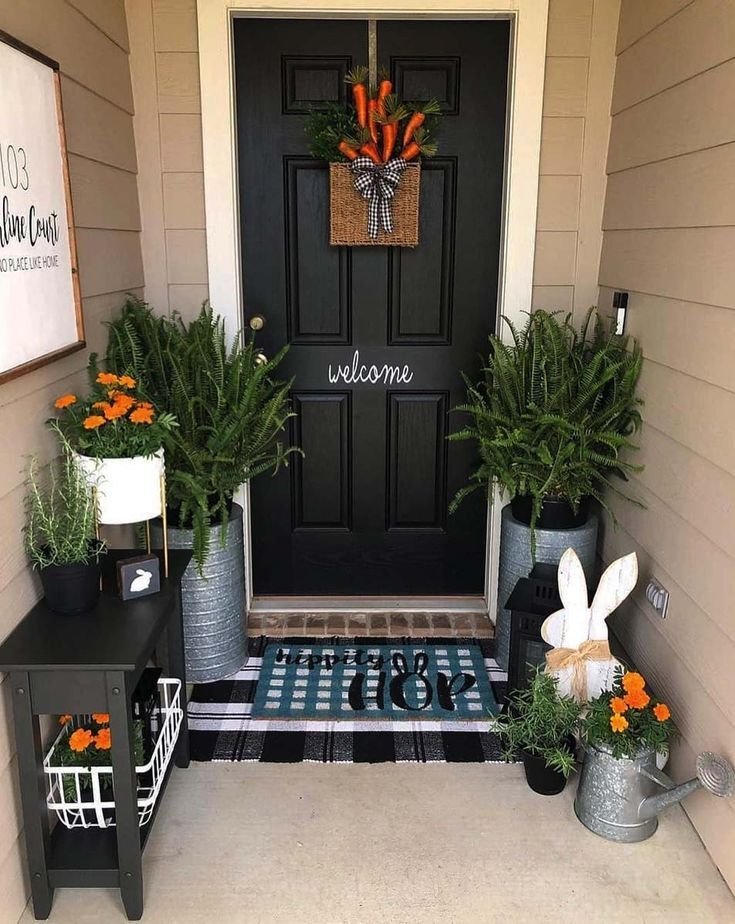 A black front door with a woven basket filled with carrots. This front porch decor is made of plants, orange flowers, rabbits and black and silver furniture.