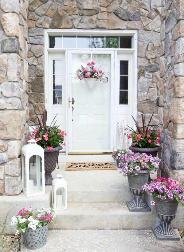 A white front door with a white basket filled with pink flowers. Antique flower pots with white lamps are placed on the steps.