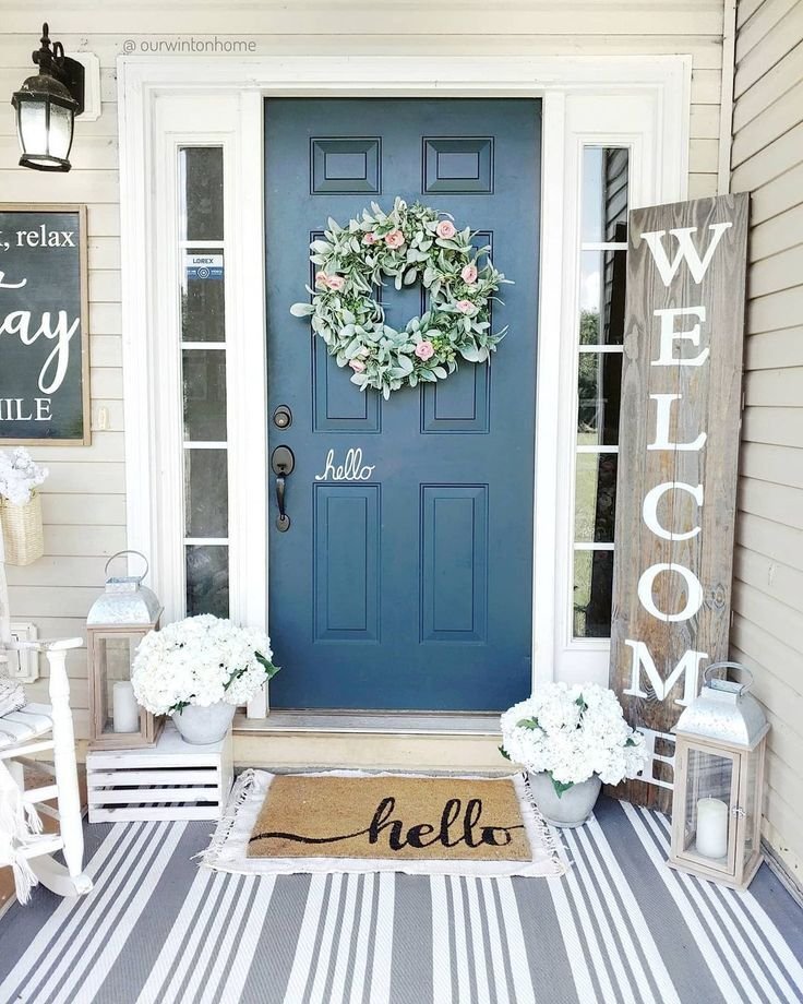 A navy front door with a summer wreath made of green leaves. A welcome wooden sign and white flowers are placed next to it.