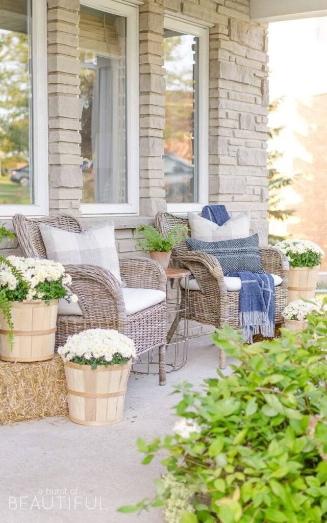 A porch decor with woven chairs, white cushions, haystacks and wooden buckets as plant pots.