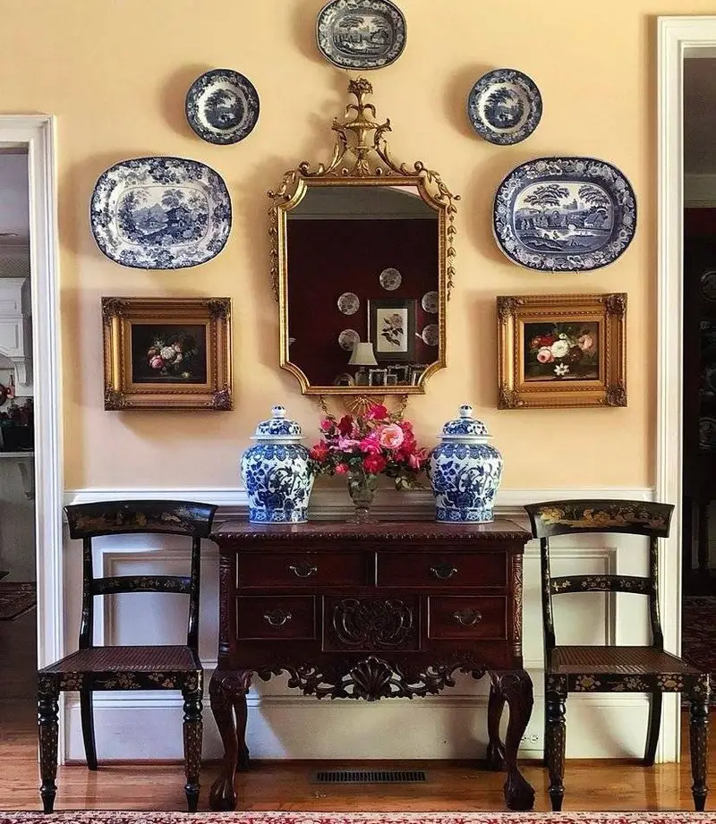 Antique plates with golden frames and vintage paintings above a vintage cabinet and next to a golden mirror.