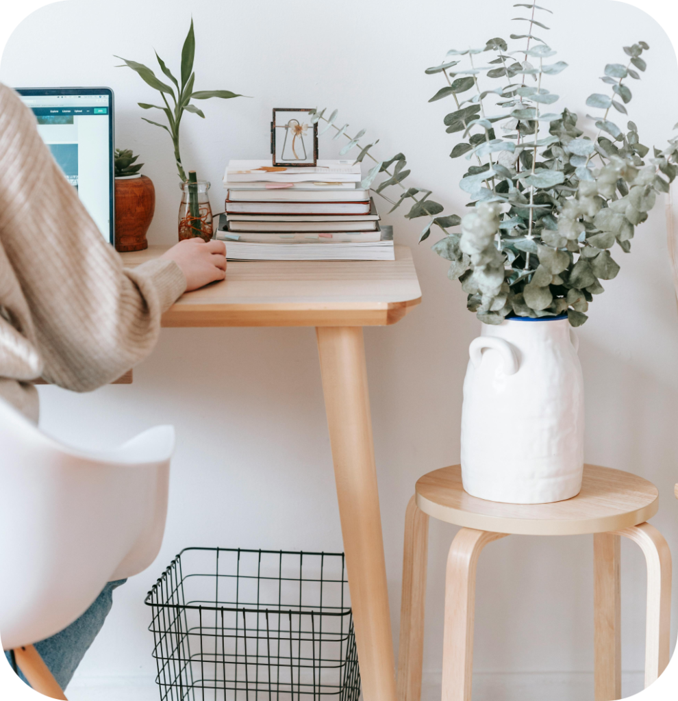 A woman working on a computer that sits on a wooden desk with plants and books.