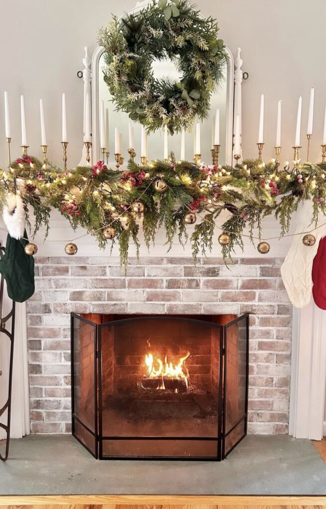 A mantel decorated with a pine garland, string lights, bells and candles.