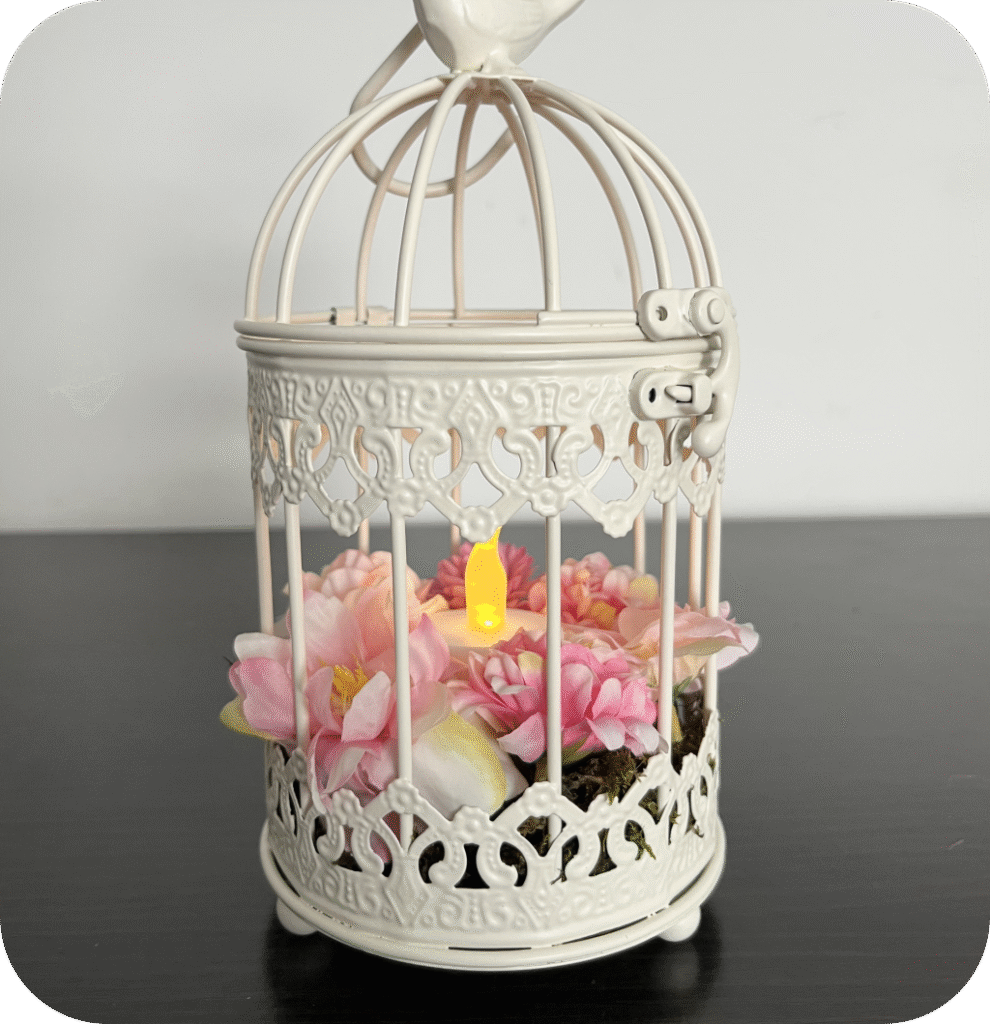 A white decorative brid cage with moss, pink flowers, and a LED candle.