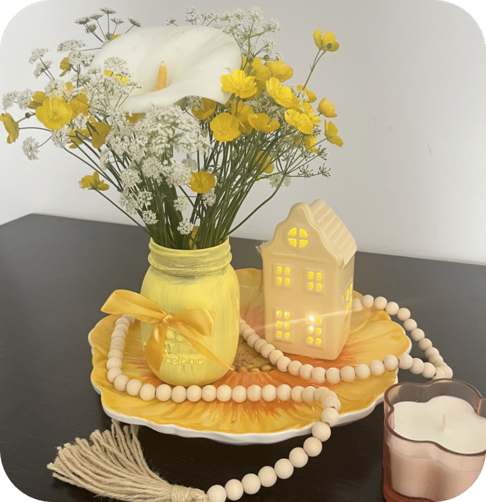 A yellow mason jar on a floral plate with a yellow house and a wooden bead garland.