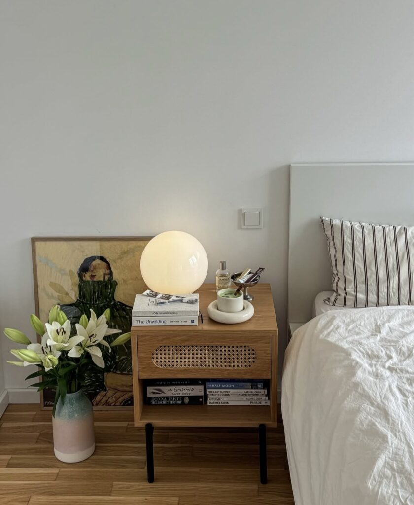 A minimalist bedroom with a cane nightstand, a painting, and lilies.