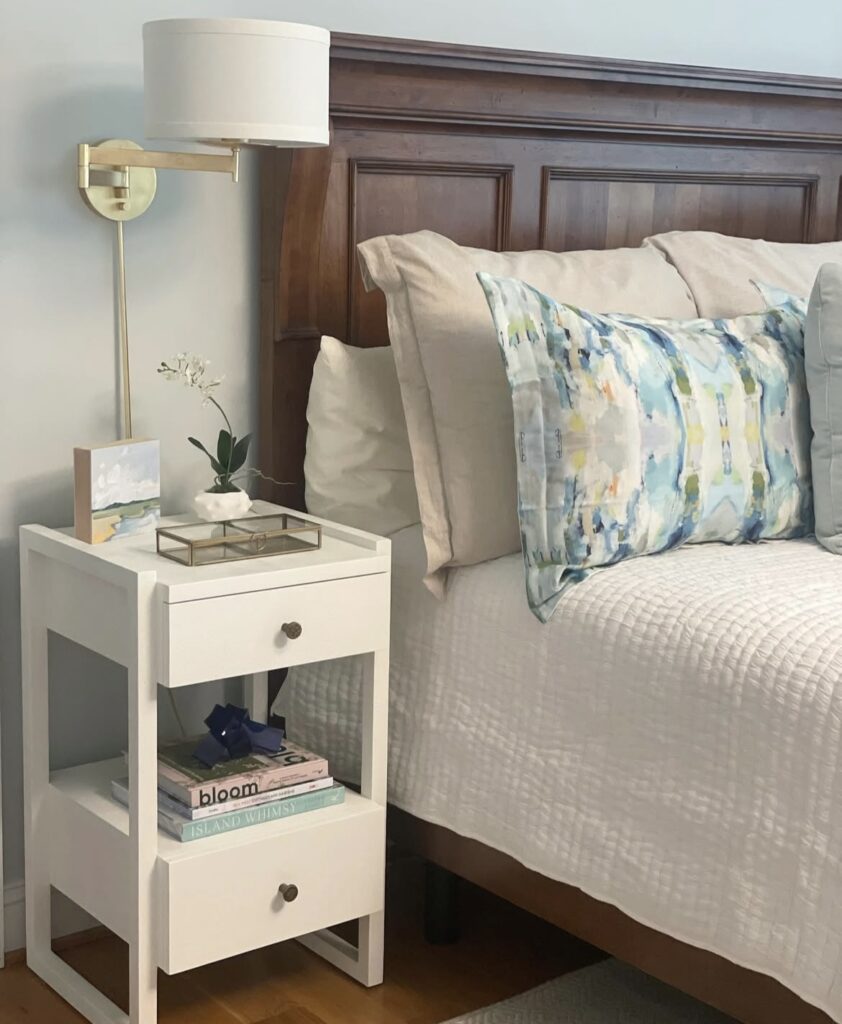 A white nightstand with books and a seascape canvas.